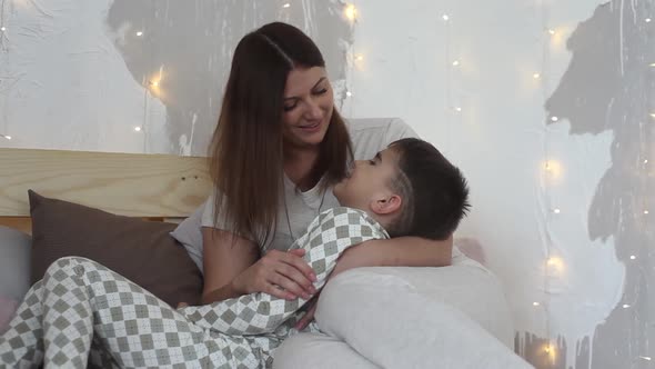 A Beautiful Mother Plays with Her Son Sitting on the Bed and Kisses Him Having a Good Mood alt