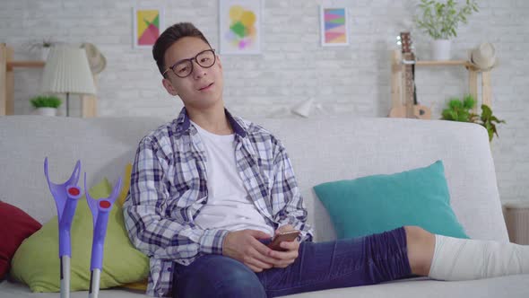 Cheerful Asian Young Man with a Broken Leg in Bandages Uses a Smartphone Looking at Camera and alt