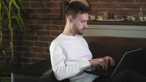 Young attractive man sitting on sofa in a cozy home and typing on a laptop alt