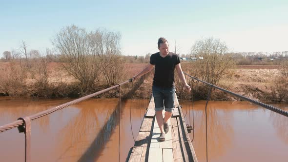 Young Man Is Walking on a Suspended Wooden Bridge Over the River.