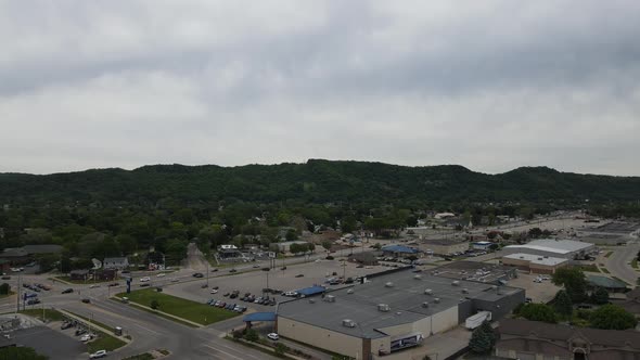 Aerial view of city edge with businesses lining highway, forest covered mountains with cloudy sky.