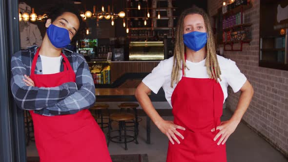Two diverse male baristas wearing face masks and aprons standing in doorway of cafe alt
