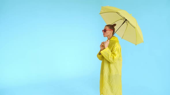 A Happy Woman with Yellow Umbrella and Yellow Raincoat Stands on Blue Background