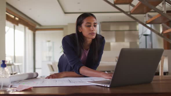 Mixed race woman by desk at home using laptop looking through blueprints alt