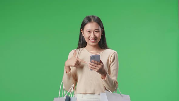 Asian Shopping Woman With Shopping Bags Pointing Mobile Phone And Smiling At Green Screen Studio alt