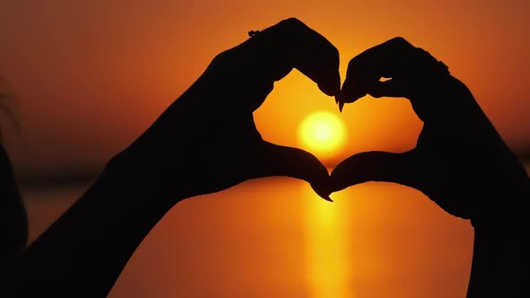 Silhouette of Female Hands Holding Sign in the Shape of a Heart at Orange Sunset alt
