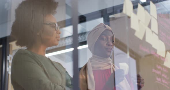 Two smiling african american businesswomen brainstorming using memo notes on glass wall in office alt
