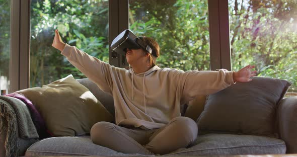 Asian girl smiling and gesturing while wearing vr headset sitting on the couch at home alt