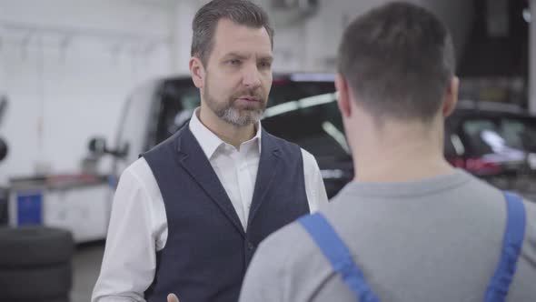 Close-up Portrait of Displeased Caucasian Man Talking with Unrecognizable Auto Mechanic. Adult alt