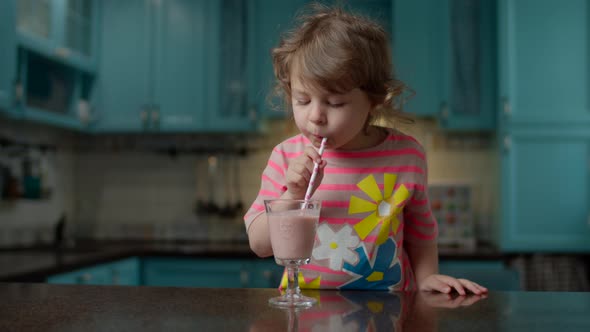 Cute little girl in pink t-shirt drinking berry smoothie with paper straw at home alt