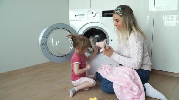 Mother with Little Smiling Daughter Sit on Floor and Put Soft Toys for Washing