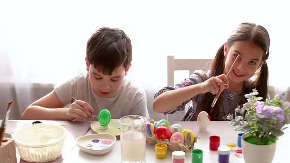 Two Children in a Room By the Window are Sitting at a White Table Painting Eggs