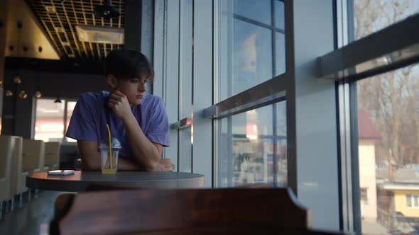 A Young Guy is Sitting in a Cafe By the Big Window Drinking Juice