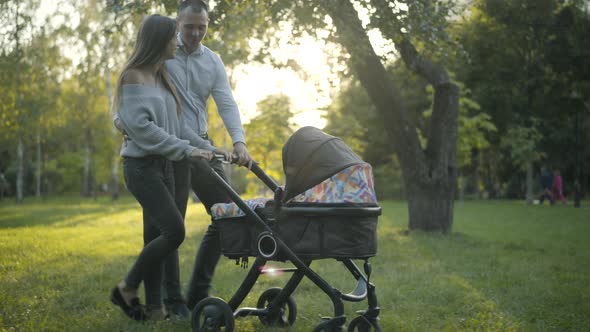 Side View Wide Shot of Cheerful Young Couple of Parents Walking with Baby Carriage in Sunlight and alt
