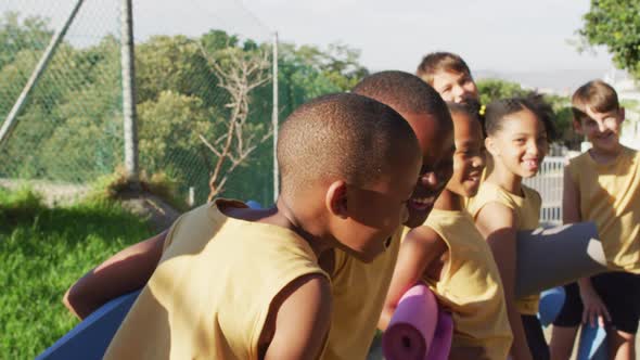 Diverse group of schoolchildren standing holding mats before yoga lesson outdoors alt