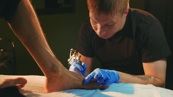 Tattooer in Gloves with Retro Coil Machine Paints Clients Foot with Ink Outline