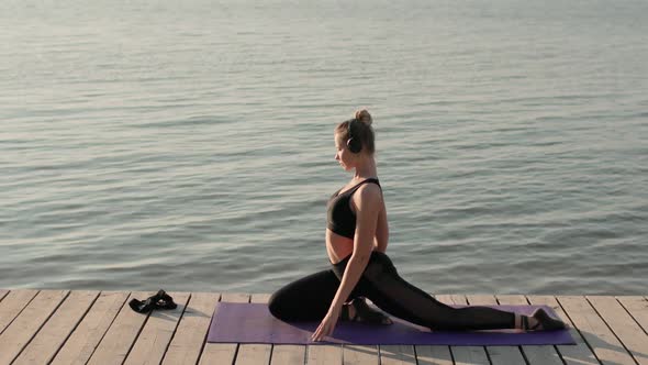 Woman Is Practicing Yoga and Doing Pigeon Pose on Mat on River Pier, Side View.