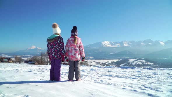 Back View of Children Standing on Rural Roadway in Snow Looking at Mountain Range in Sunshine
