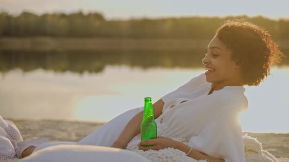 Beautiful Black Girl Chatting and Drinking While Laying on a Beach at the Sunset
