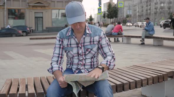 Caucasian Man Sitting on a Bench in a Square in the City Studying a Map of City alt