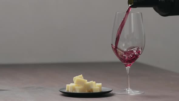 Slow Motion Pour Red Wine Into Wineglass with Hard Cheese on Background on Wood Table alt