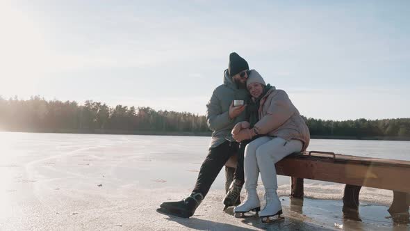 Cute Couple Drink Hot Tea From Thermos and Kiss Sitting on a Dock in Winter alt