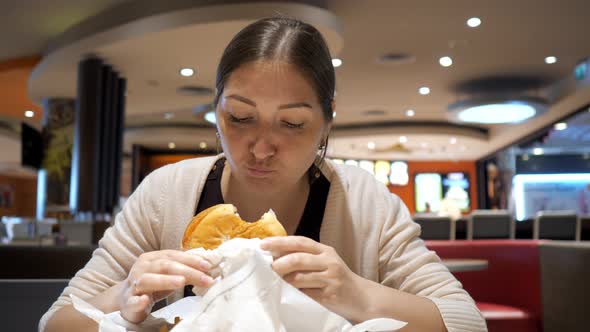 Young Brunette Woman is Eating Burger in Fast Food Restaurant in Food Court alt