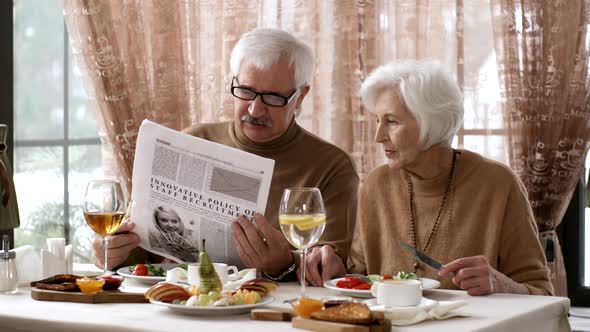 Senior Lady Eating Breakfast at Hotel and Husband Reading Newspaper alt
