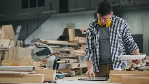Man Reading Document Working with Tablet in Wood Workshop Touching Screen alt