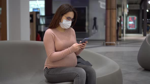 A Young Pregnant Woman in Glasses and a Medical Mask Sits with a Phone on a Bench alt