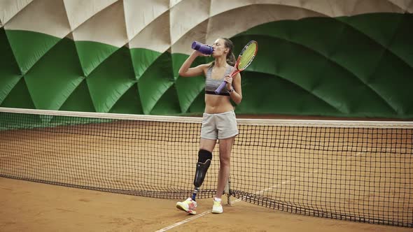 Seductive Tennis Girl with the Leg Prosthesis is Standing Near the Net on the Tennis Court Drinking alt