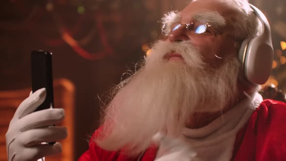 Cheerful Old Santa Claus Is Enjoying By Music From Headphones and Smartphone at Christmas Holidays alt