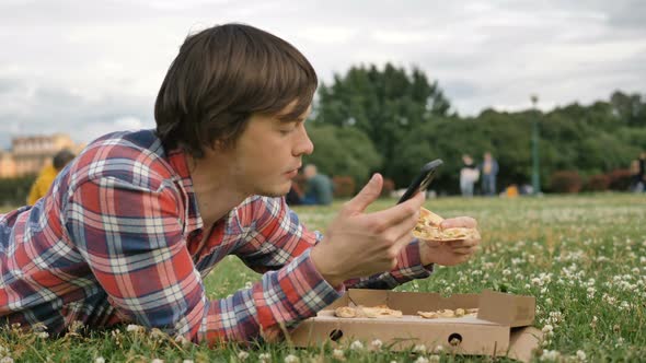 Man Lying on the Grass Eating Pizza and Using a Smartphone in a City Park on Nature alt