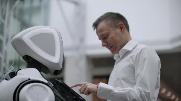 A Man Stands with a Robot Bot and Asks Him Questions and Asks for Help By Clicking on the Screen on alt
