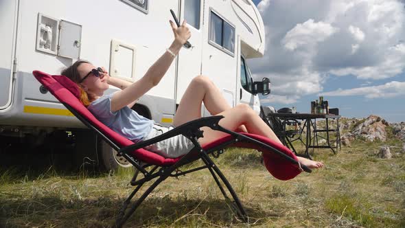 Woman Sitting Next To Camper Rv On Folding Chair Making Selfie By Phone