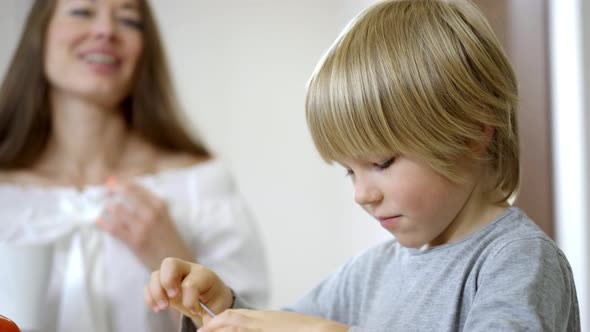 Side View Closeup of Blond Cute Boy Eating Healthful Morning Cereals with Blurred Woman Admiring Son alt