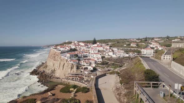Establishing shot capturing cars visiting Azenhas do Mar town with foamy waves crashing the shore. alt