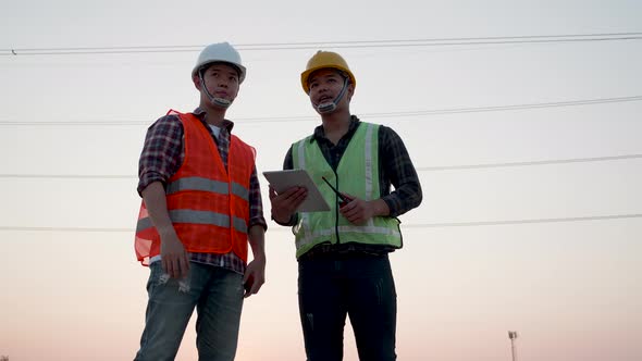 Two Asian engineers inspected by tablet at high voltage power line pylon at sunset