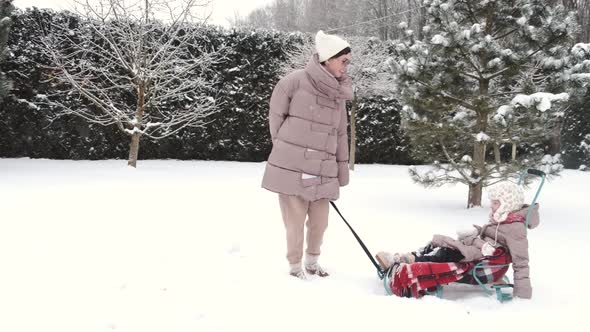 Young Happy Mother and Her Daughter Enjoying a Sledge Ride in a Beautiful Snowy Backyard alt