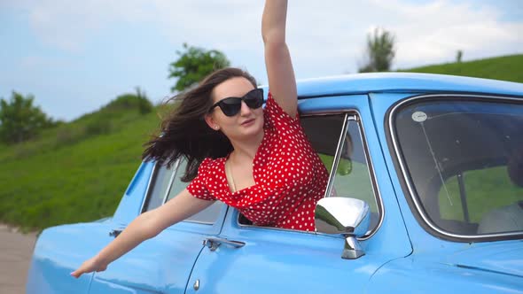 Young Girl in Sunglasses Leaning Out of Vintage Car Window and Enjoying Trip alt