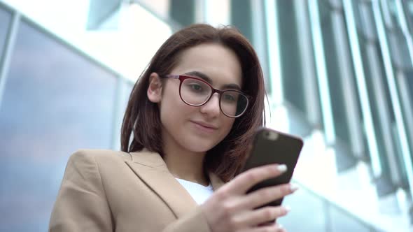 A Young Woman Stands with a Phone on the Background of a Business Center. The Girl in the Coat Is alt