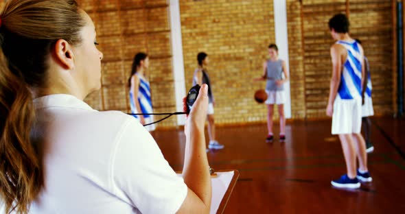 Female coach looking at stopwatch while students playing in basketball court alt