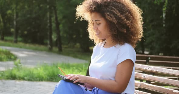 Thoughtful Black Young Woman with Curly Hair Looks Around and Writes Into Paper Notebook alt