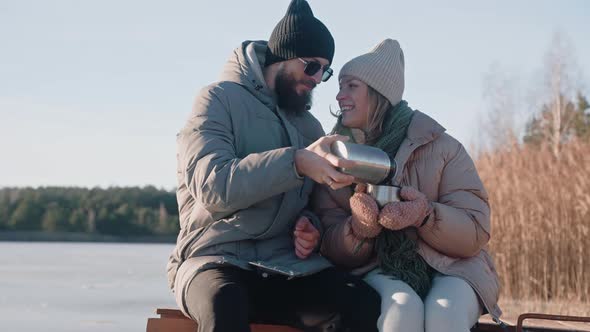 Cute Couple Drinking Hot Tea From Thermos Sitting on a Dock in Winter Near Lake