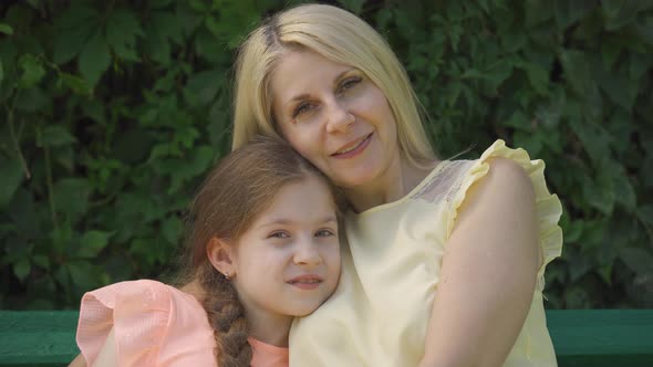 Portrait of Mature Blond Mother in Glasses and Her Little Daughter Looking at Camera While Sitting alt