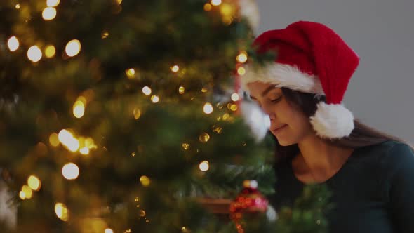 Pretty Young Female in Red Santa Hat Looking Christmas Tree Touching Ornaments alt