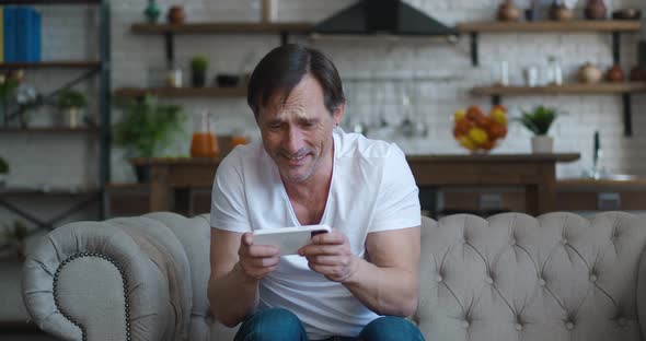 Happy Caucasian Mature Man Gamer Play Video Game Online with Smartphone App Sitting on Sofa at Home alt