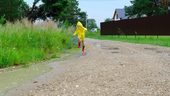 A girl in red rubber boots and a yellow raincoat runs through puddles after a rain in the village. S alt