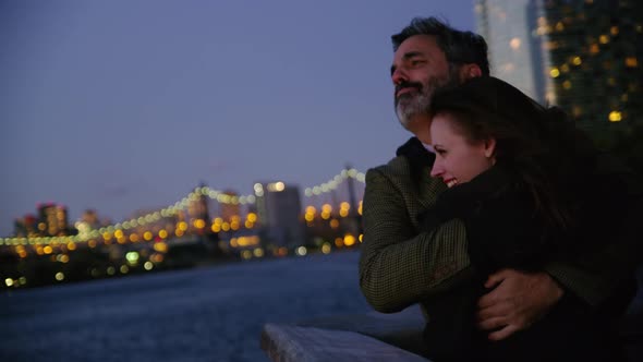 Couple in New York City standing on pier at night with city skyline in background