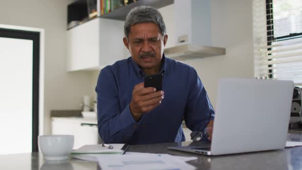 Senior mixed race man using smartphone and laptop in kitchen alt